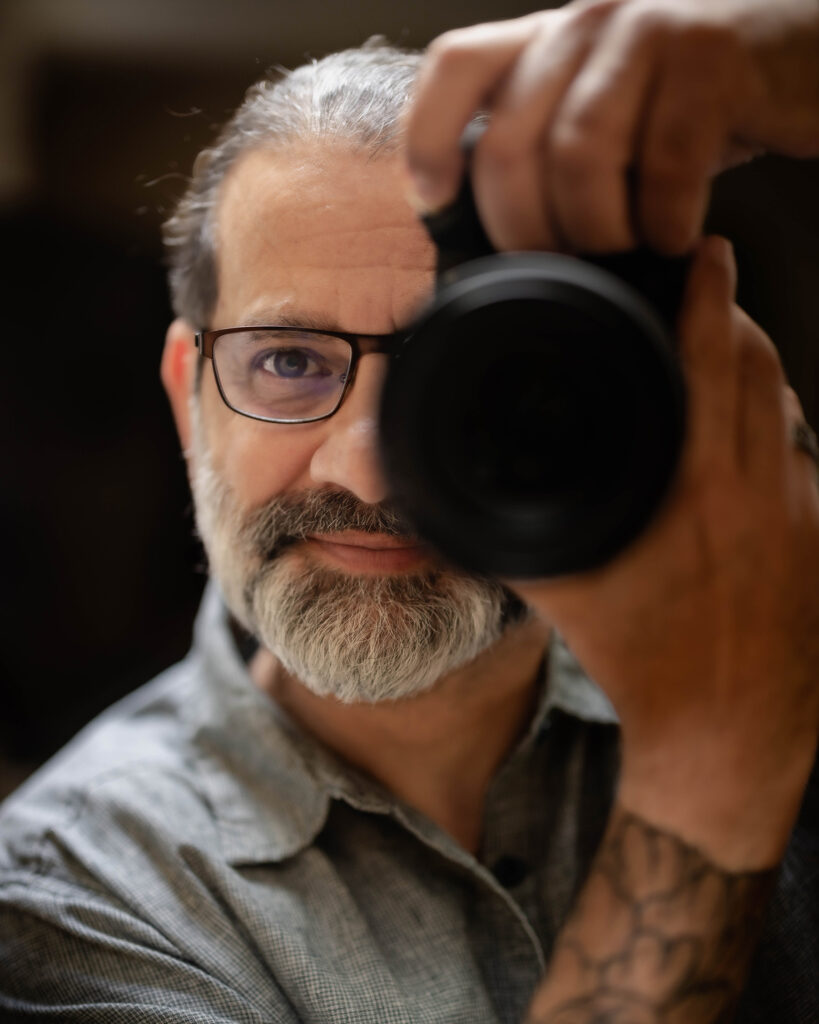 Self portrait of Teek in a grey shirt against a black studio backdrop, Ashland Oregon